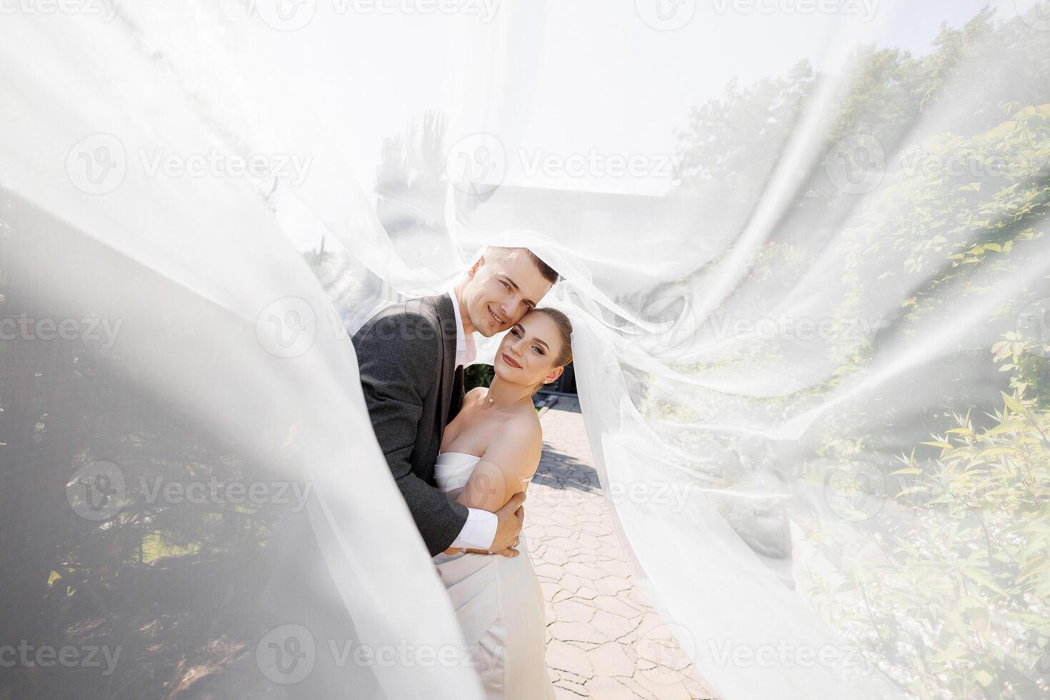 A bride and groom are posing for a picture under a white veil. The bride is wearing a white dress and the groom is wearing a gray suit. Scene is happy and romantic photo