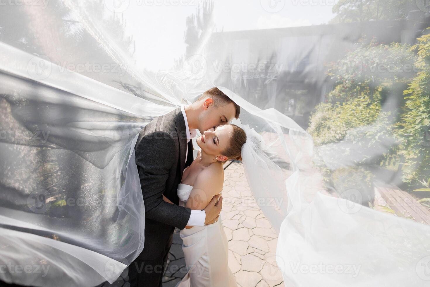 A bride and groom are embracing under a white veil. The bride is wearing a white dress and the groom is wearing a suit. The scene is intimate and romantic, capturing the love photo