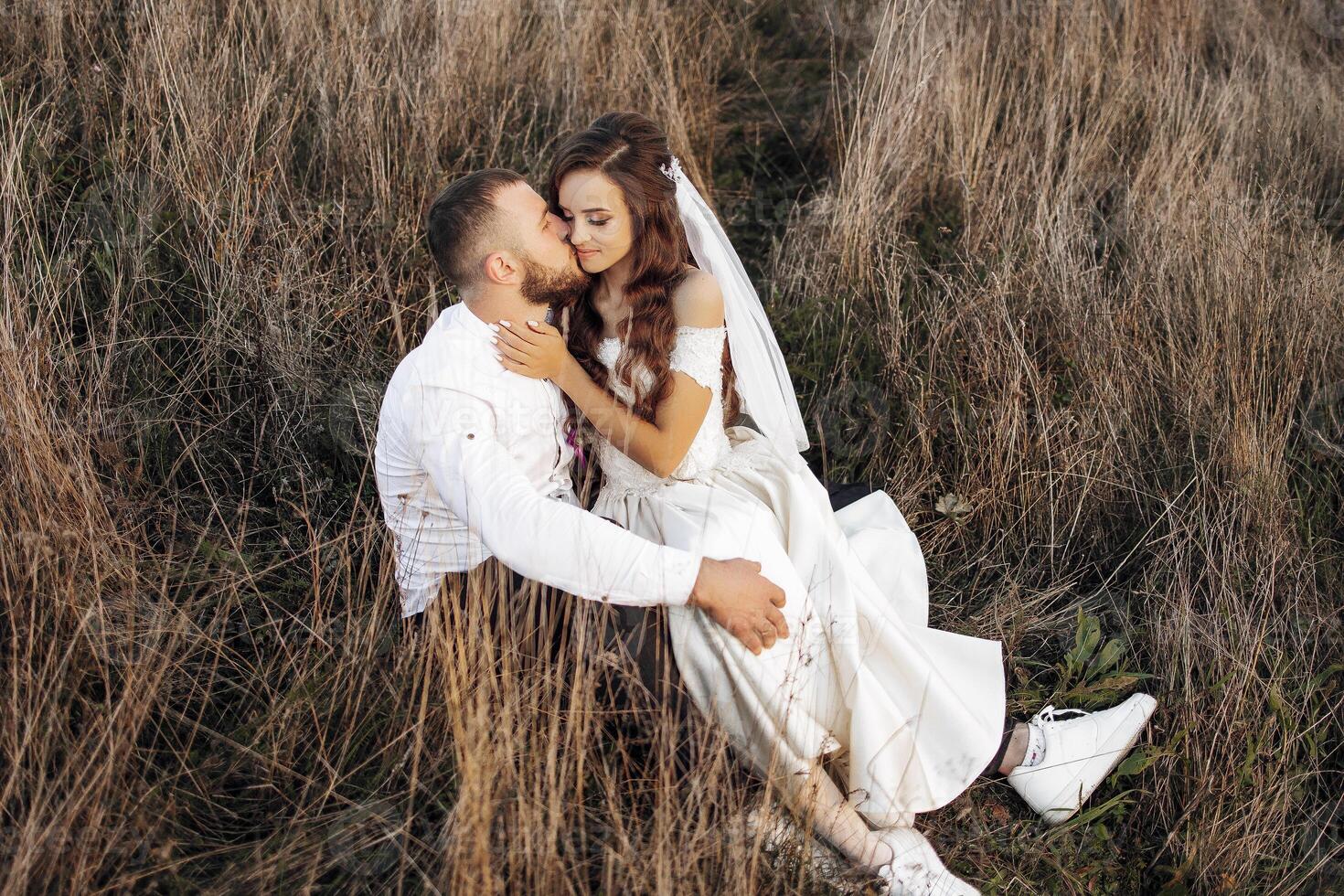 A bride and groom are sitting in a field, kissing each other. Scene is romantic and intimate photo