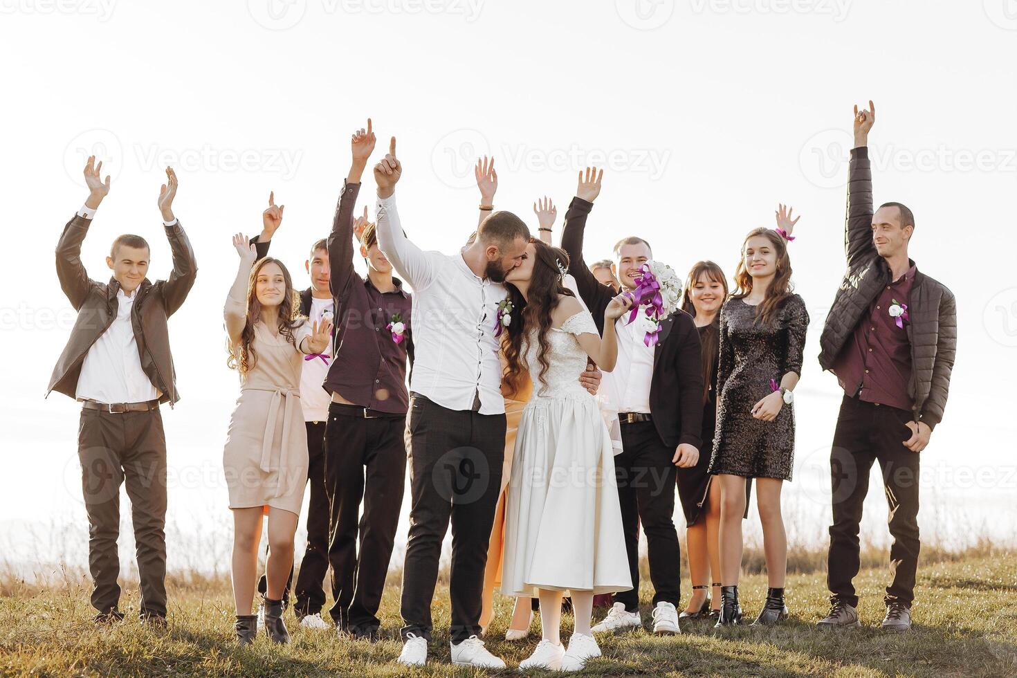 A group of people are posing for a picture, with a bride and groom in the center. The bride is wearing a white dress and the groom is wearing a suit. The group is standing on a grassy field photo