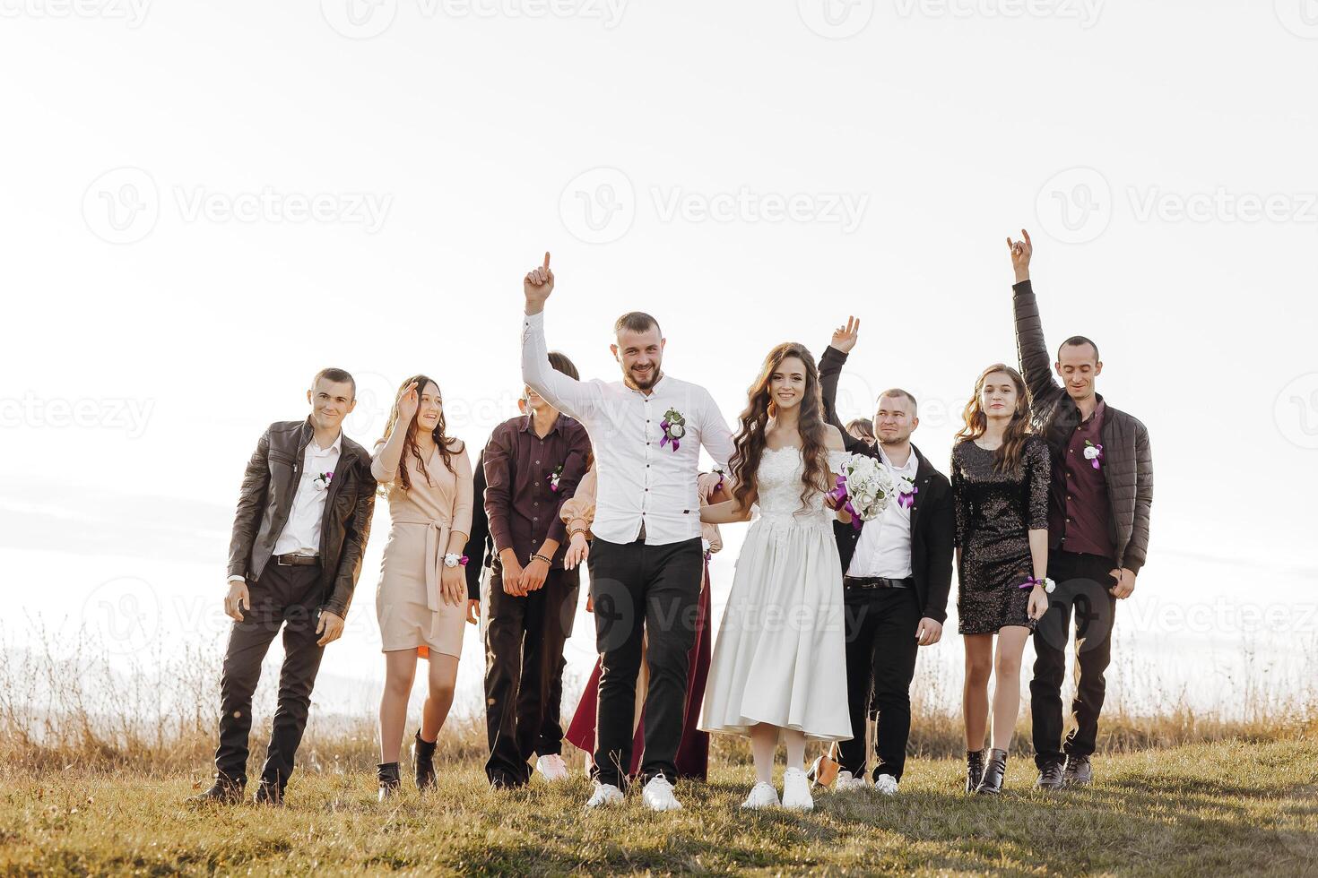 A group of people are standing in a field, with a bride and groom in the center. The bride is holding a bouquet and the groom is holding her hand. The group is all smiling and looking happy photo