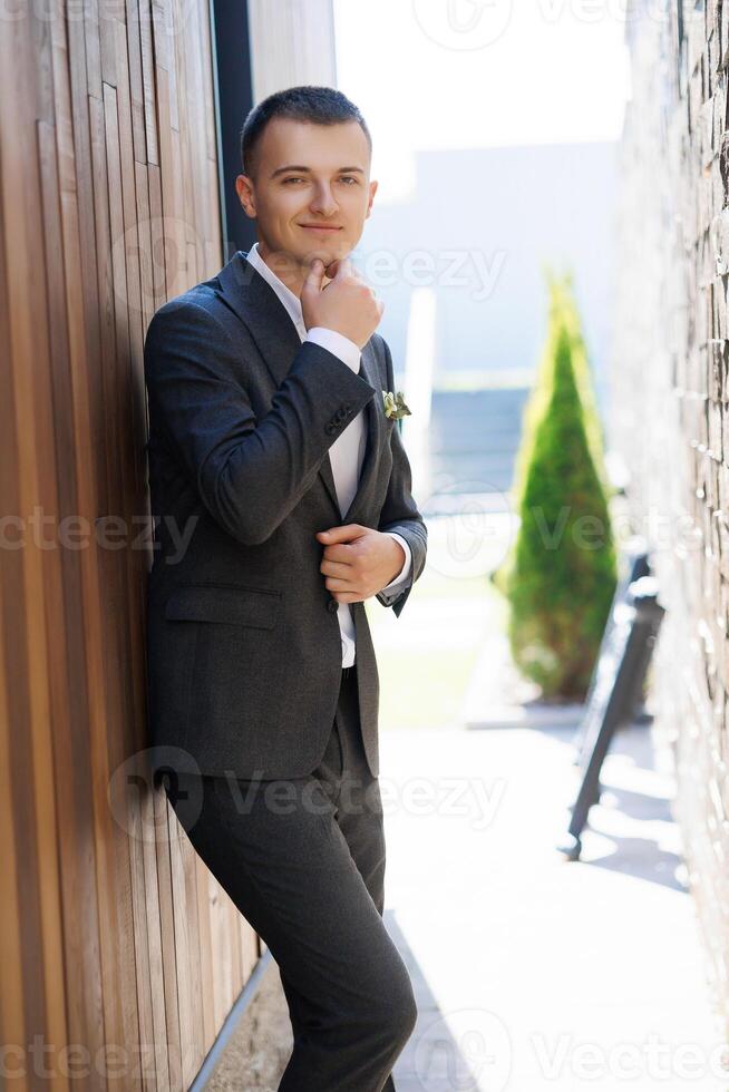 A man in a suit is leaning against a wall. He is wearing a white shirt and a green flower in his pocket photo
