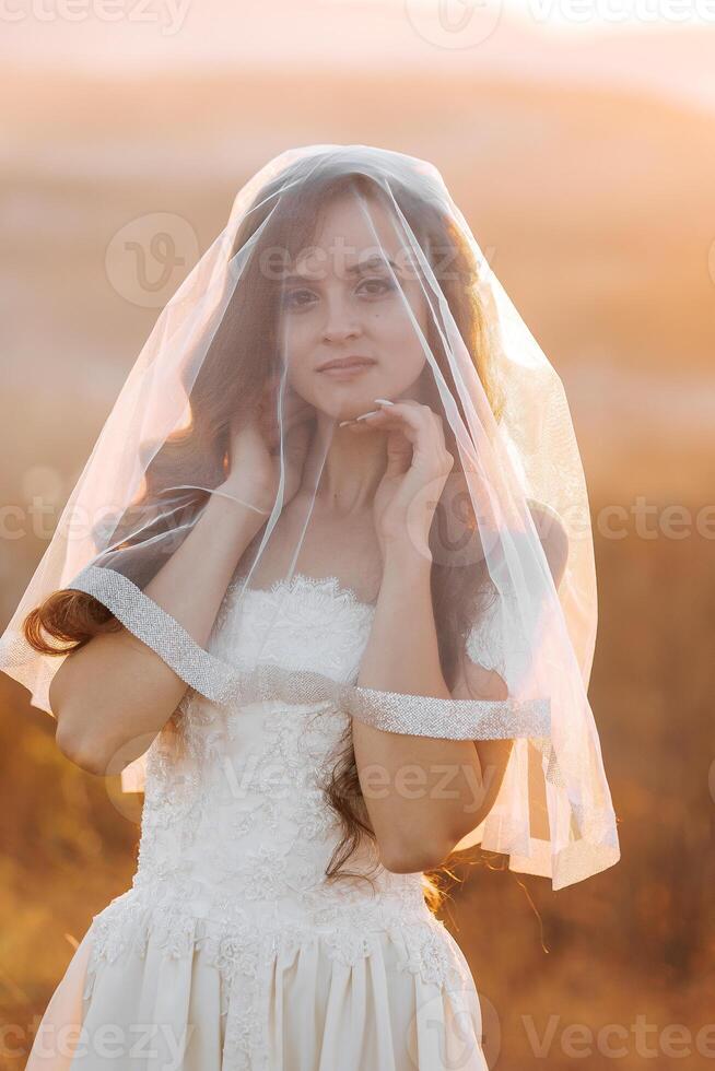 A woman in a white dress is standing in the sun with a veil covering her face. The veil is made of a net-like material and is covering her face, giving her a mysterious and ethereal appearance photo