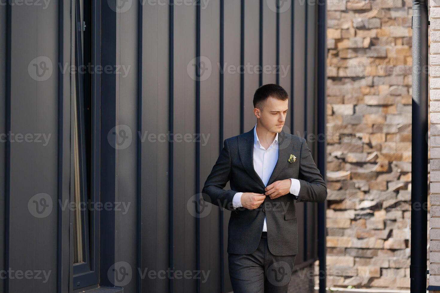 A man in a suit is standing in front of a brick wall. He is adjusting his jacket and looking at the camera photo