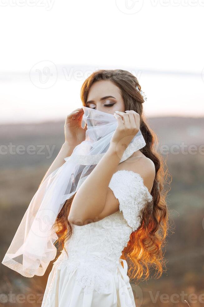 A woman in a white dress is wearing a scarf and looking away from the camera. The image has a serene and romantic mood, as the woman is in a beautiful outdoor setting photo