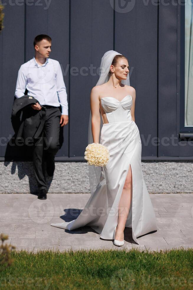 A bride and groom are standing outside in front of a building. The bride is wearing a white dress and holding a bouquet of flowers. The man is wearing a suit and tie. Scene is happy and celebratory photo