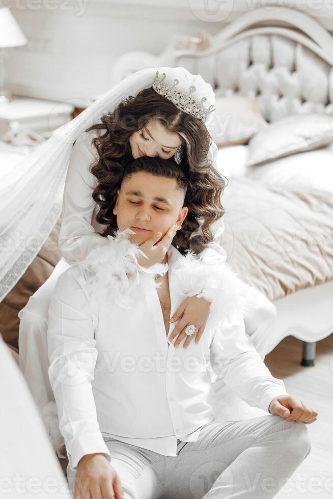 A man and woman are posing for a picture, with the woman wearing a white wedding dress and the man wearing a white shirt. The man is sitting on the bed while the woman is standing on top of him photo