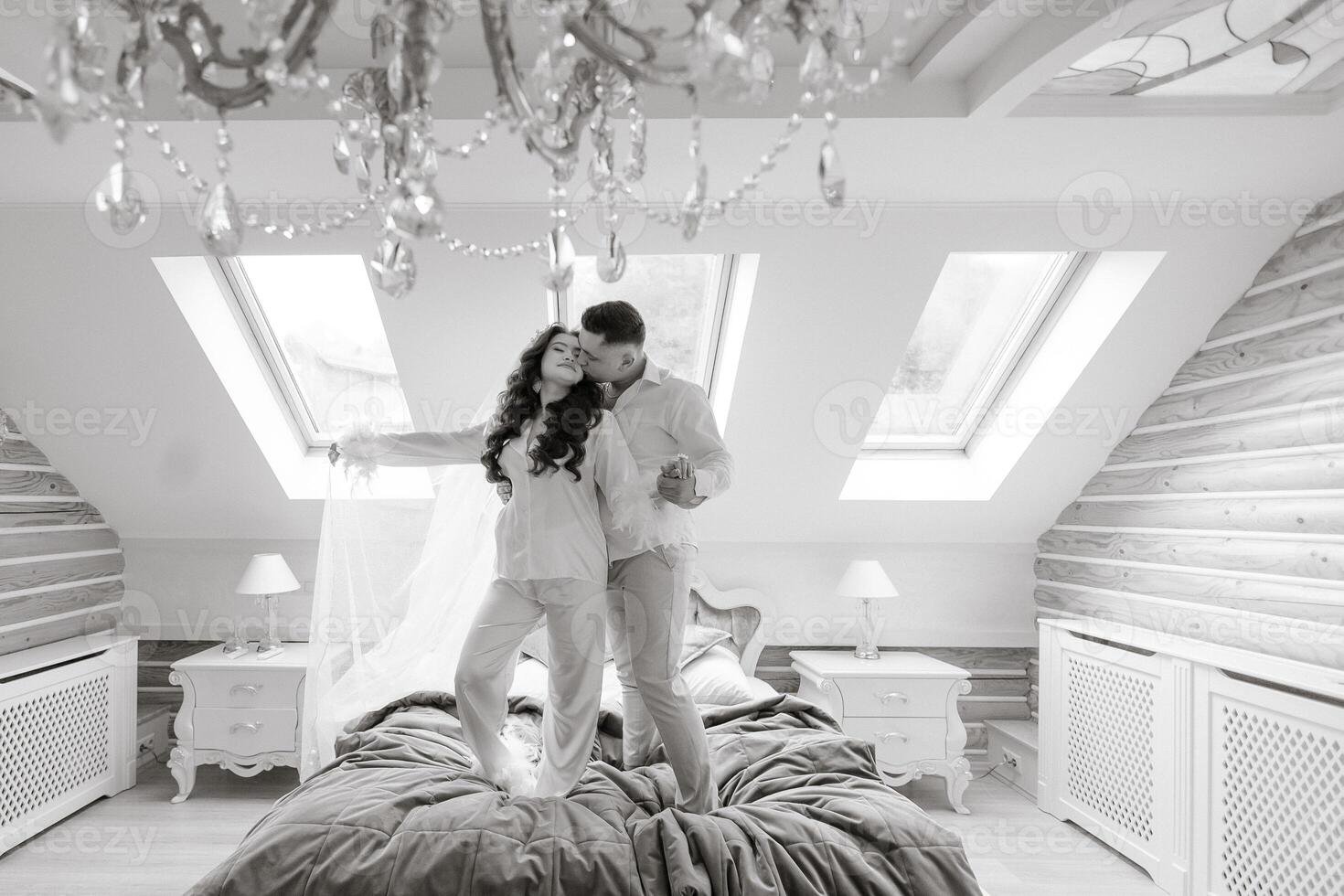 A couple is standing on a bed in a room with a chandelier above them. Scene is romantic and intimate photo