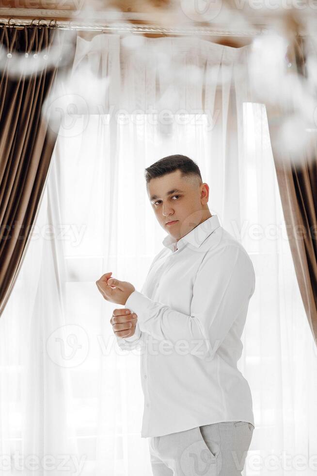 A man in a white shirt is standing in front of a window, adjusting his shirt. Concept of formality and attention to detail, as the man takes care to ensure his appearance is neat and tidy photo