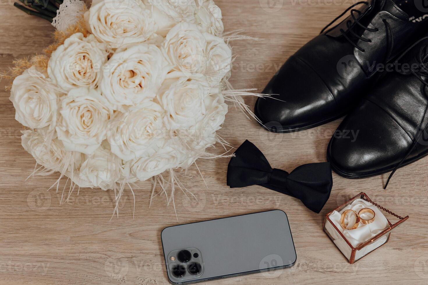 A bouquet of white roses sits on a table next to a cell phone and a pair of black shoes photo