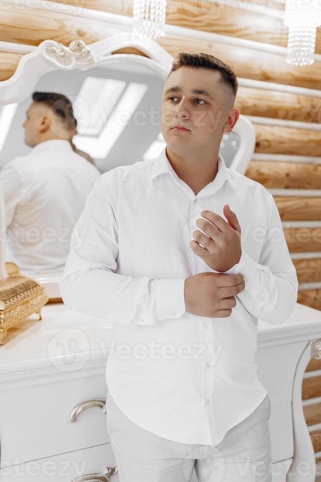 A man in a white shirt and white pants is standing in front of a mirror. He is adjusting his shirt and looking at himself in the mirror. Concept of self-care and attention to detail photo