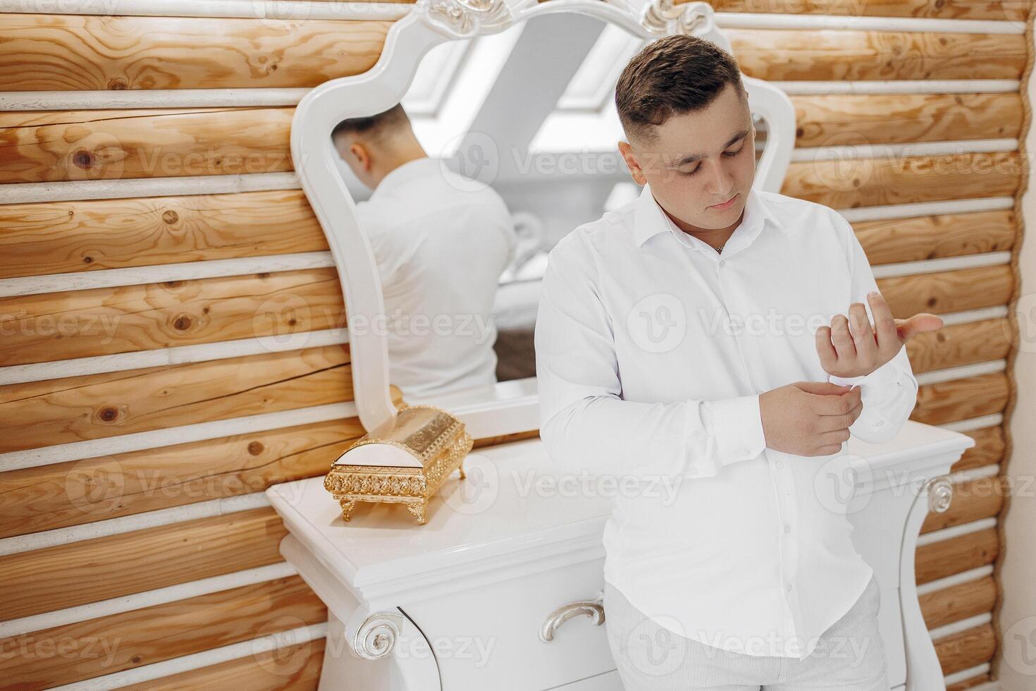A man is getting dressed in front of a mirror. He is wearing a white shirt and white pants photo