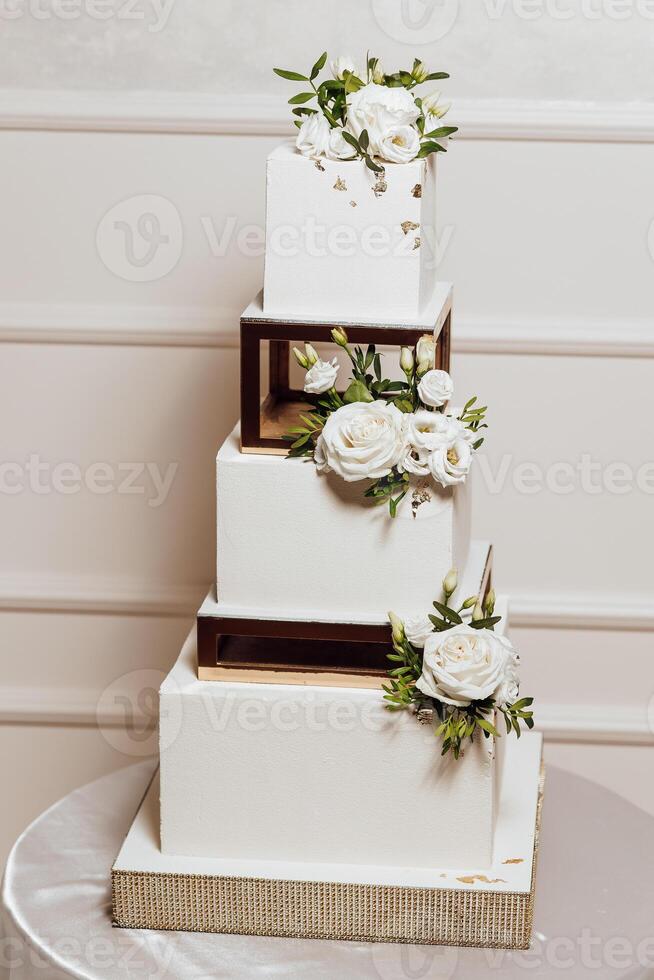 A four tier wedding cake with white flowers and gold accents. The cake is placed on a table and is the centerpiece of the room photo
