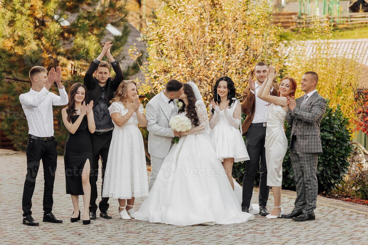 A group of people are posing for a picture, with a bride and groom in the center. The bride is wearing a white dress and the groom is wearing a suit. The group is smiling and clapping photo