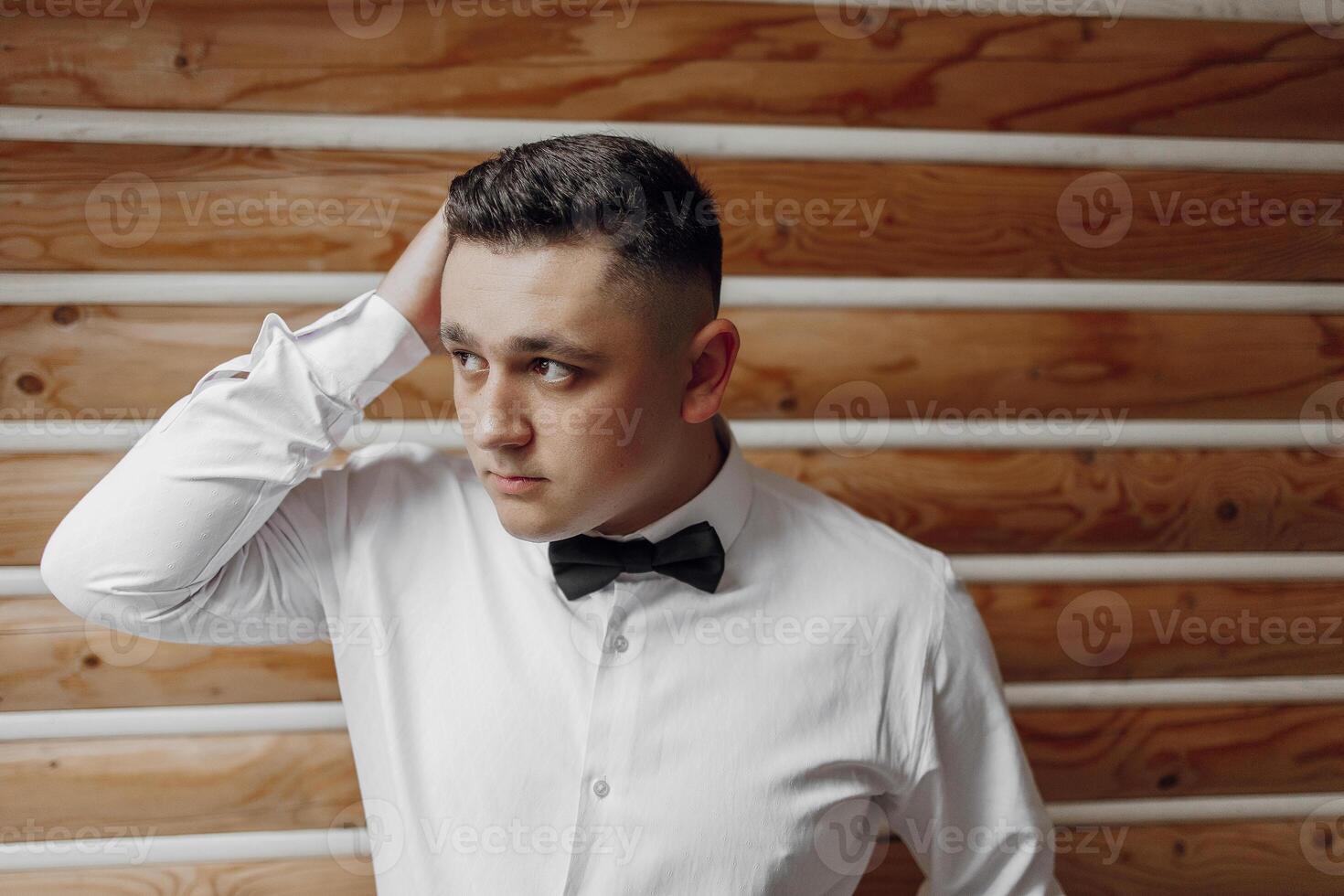 A man in a white shirt and black bow tie is standing in front of a wooden wall. He is looking at the camera with a serious expression photo