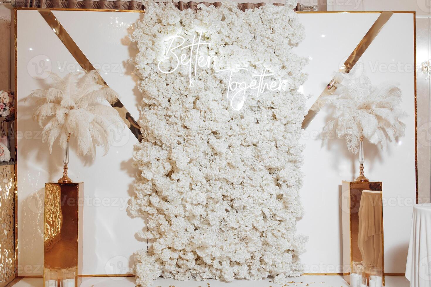 A white wall with a sign that says and a white flower wall. There are two tall white flower plants on either side of the wall photo