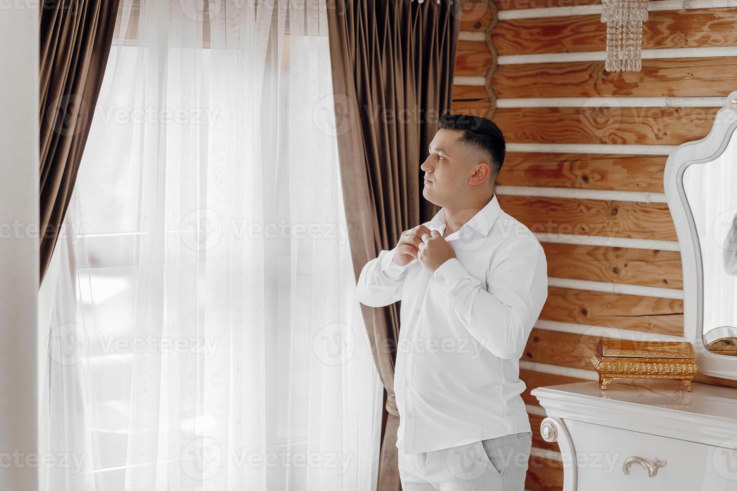 A man is getting dressed in front of a mirror. He is wearing a white shirt and gray pants photo