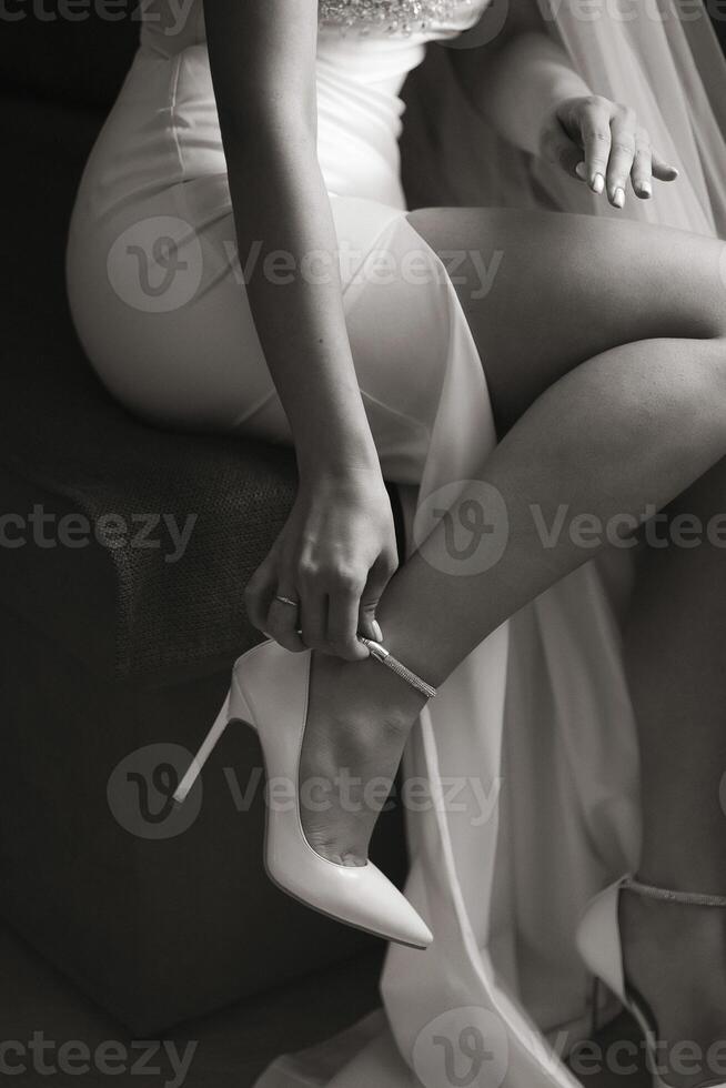 A woman in a white dress is sitting on a couch and putting on her wedding shoes. Concept of elegance and sophistication, as the woman is dressed in a wedding gown and preparing for a special occasion photo