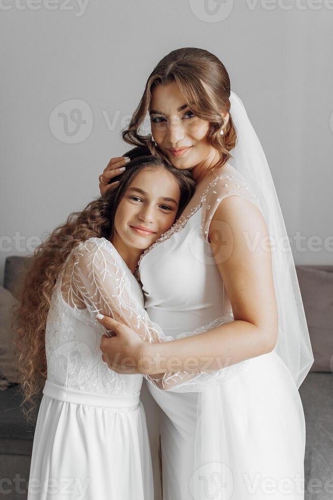 A woman and a little girl are hugging each other. The woman is wearing a wedding dress and the girl is wearing a dress. Scene is warm and loving photo