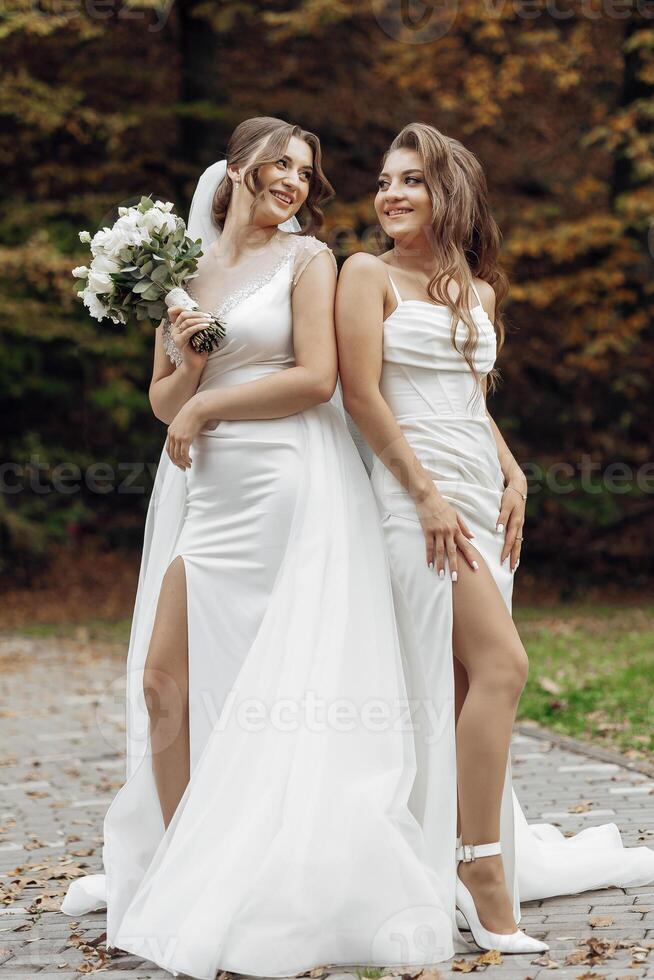 Two women in white wedding dresses pose for a picture. One of them is holding a bouquet of flowers photo