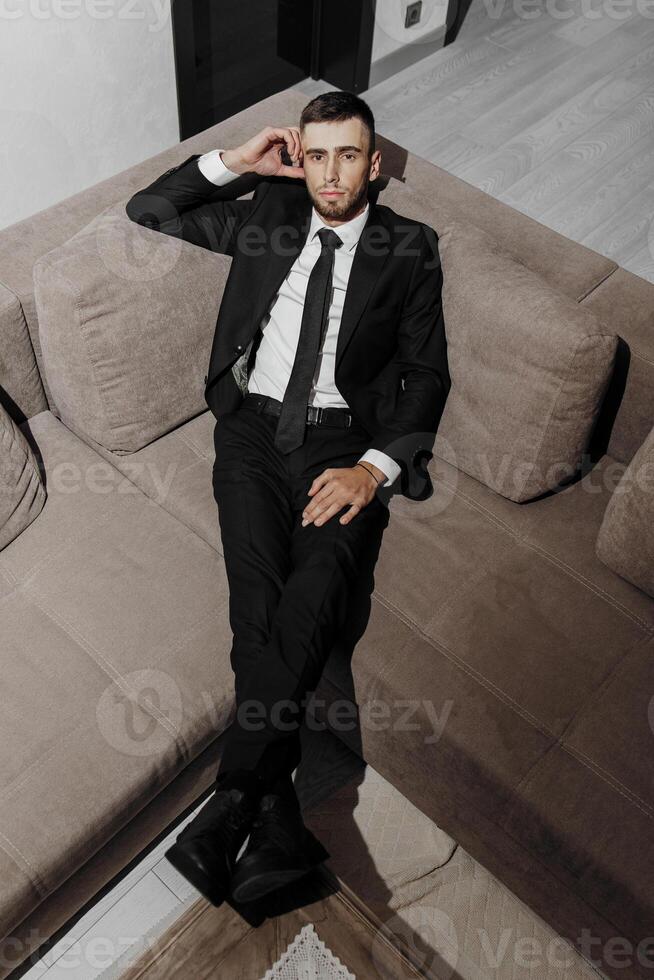 A man in a suit and tie is sitting on a couch. He is wearing a black suit and a white shirt photo