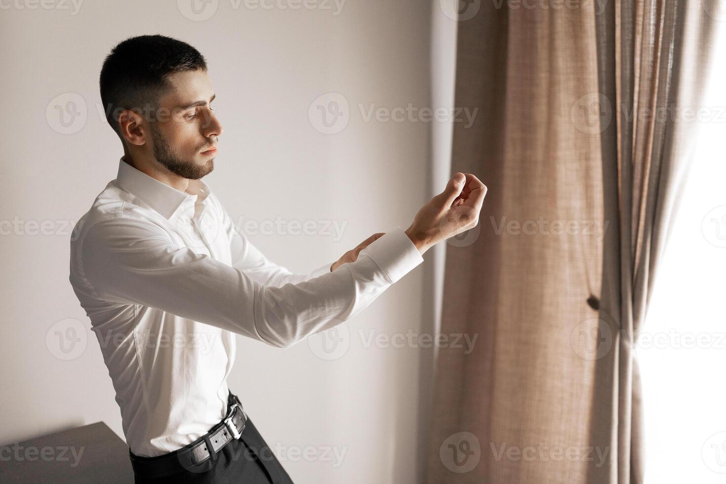 A man in a white shirt and black pants is standing in front of a curtain. He is holding his arm out and he is in a contemplative or relaxed mood photo