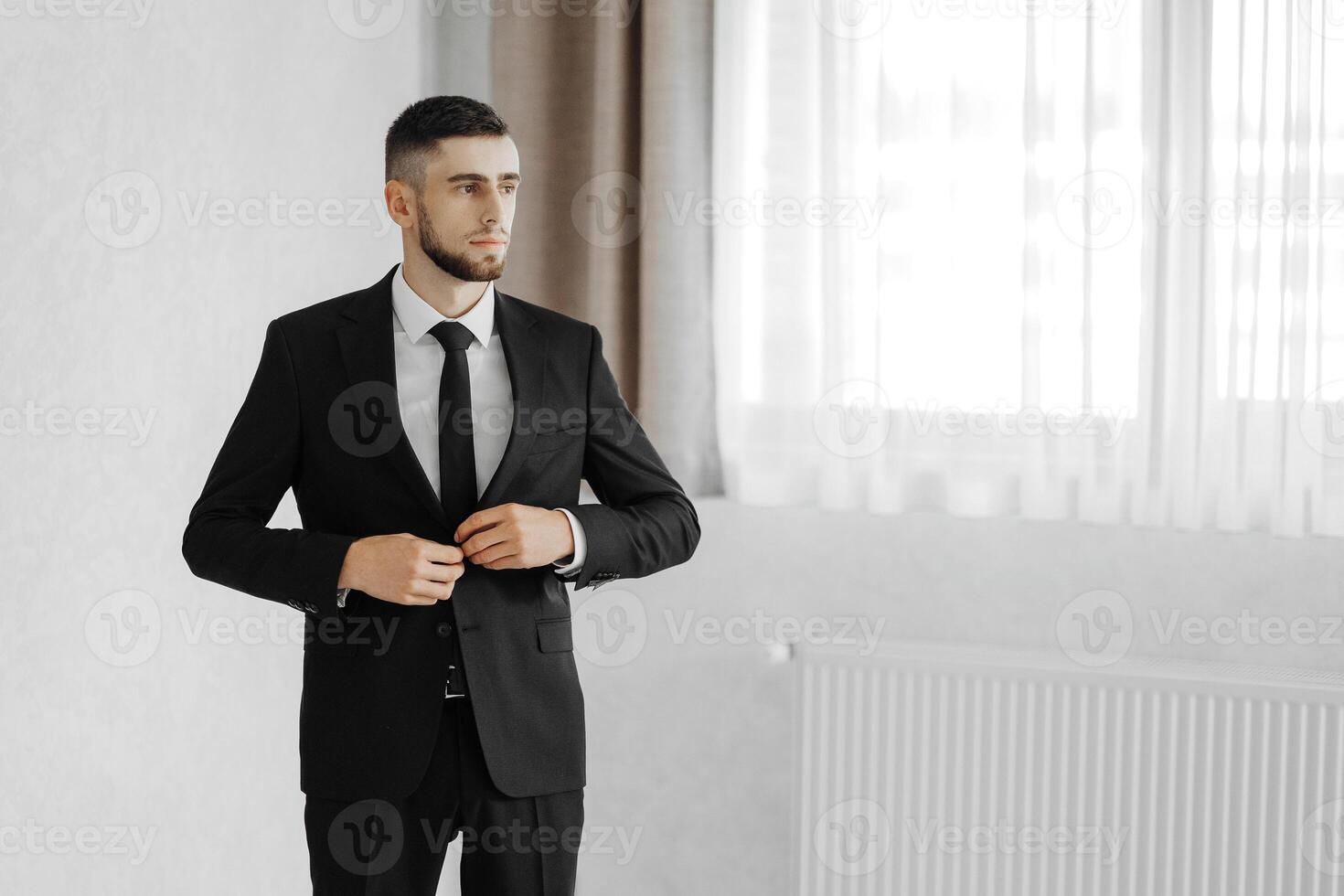 A man in a suit and tie is standing in front of a window. He is adjusting his tie and looking out the window. Concept of formality and professionalism, as the man is dressed in a suit and tie photo