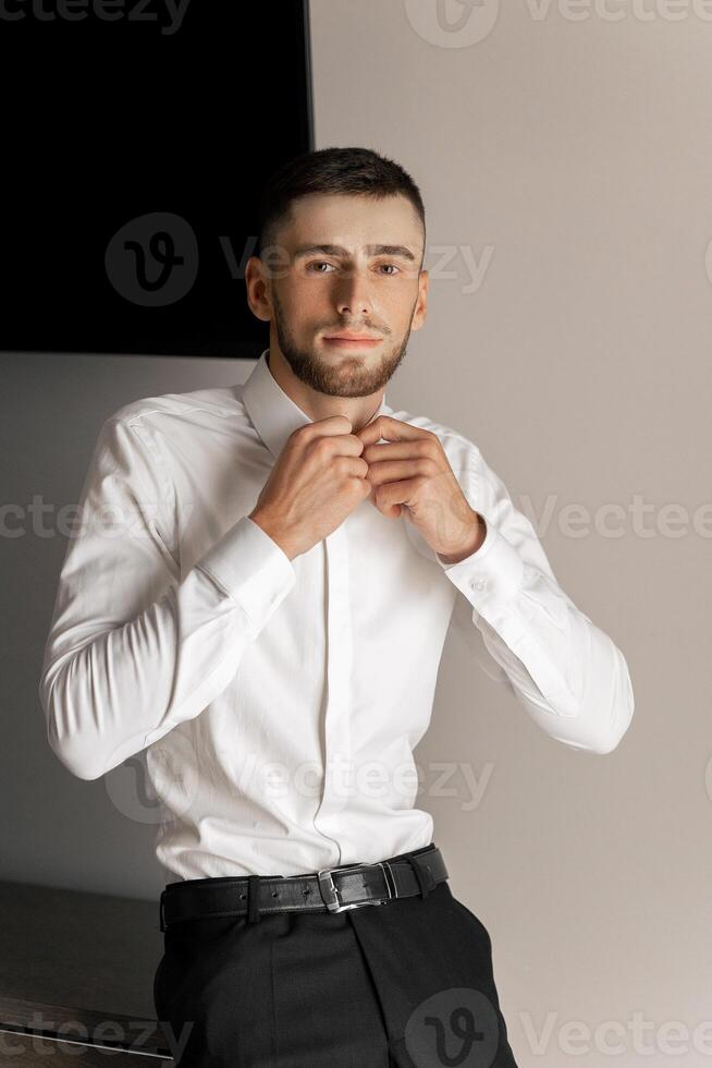 A man in a white shirt and black pants is standing in front of a wall. He is adjusting his shirt and looking at the camera. The image has a casual and relaxed mood photo
