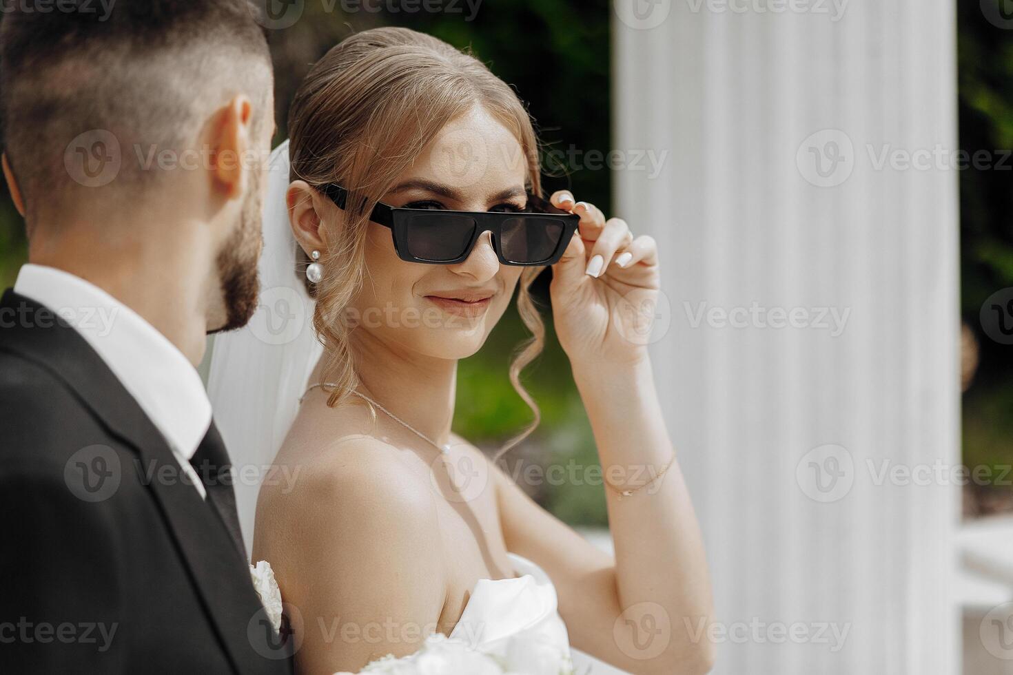 A bride and groom are posing for a picture with sunglasses on. The bride is wearing a white dress and the groom is wearing a black suit. The sunglasses add a cool and stylish touch to the photo