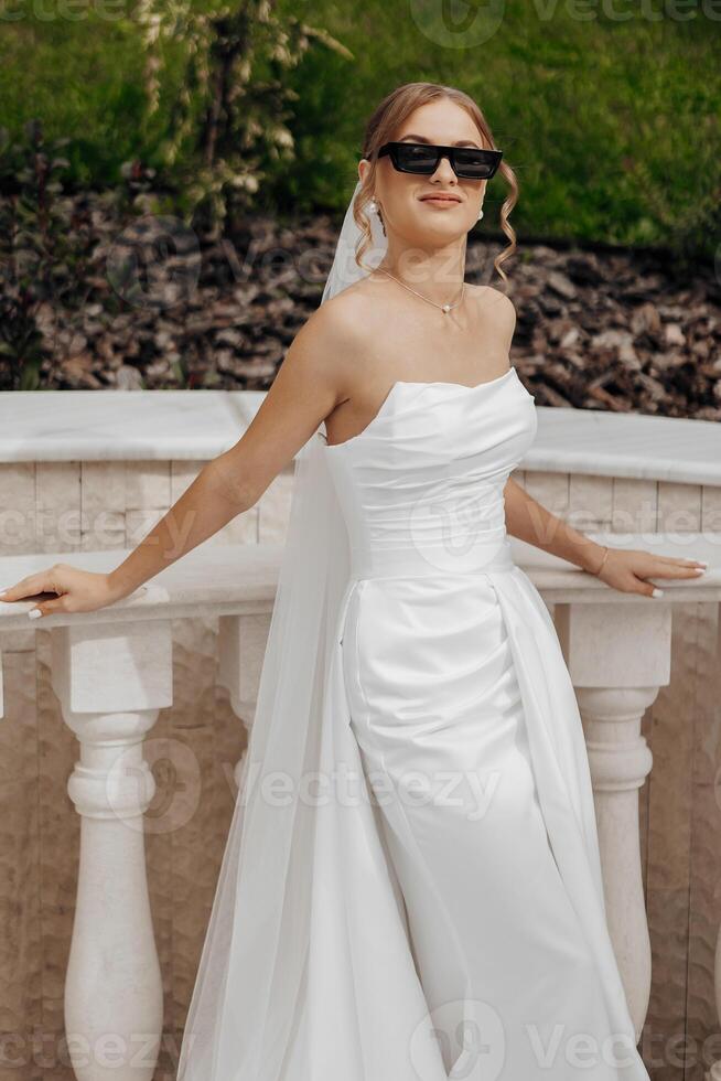 A woman in a white dress is standing on a balcony, wearing sunglasses and a white veil. She is smiling and she is enjoying the view photo