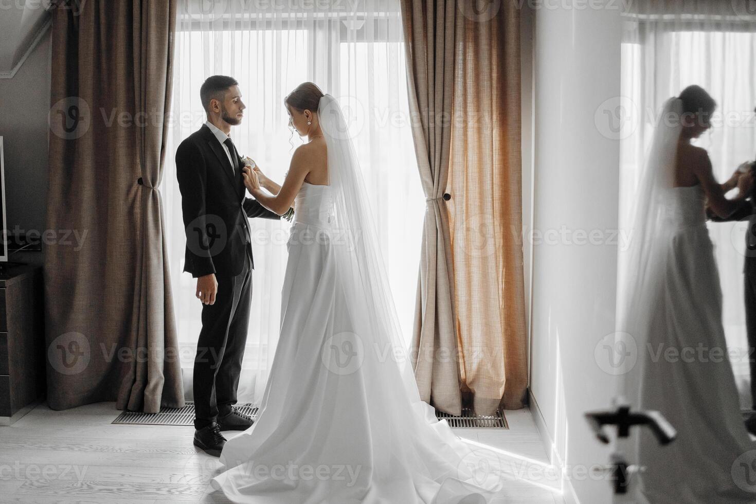 A bride and groom are getting ready for their wedding. The bride is wearing a white dress and the groom is wearing a suit. They are standing in front of a window with curtains photo