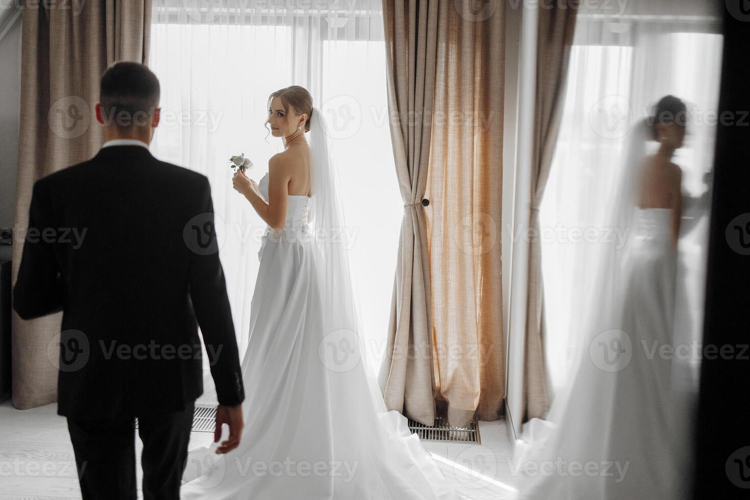 A bride and groom are standing in front of a window with a bride holding a bouquet. The man is wearing a suit and the woman is wearing a white dress. Scene is romantic and happy photo