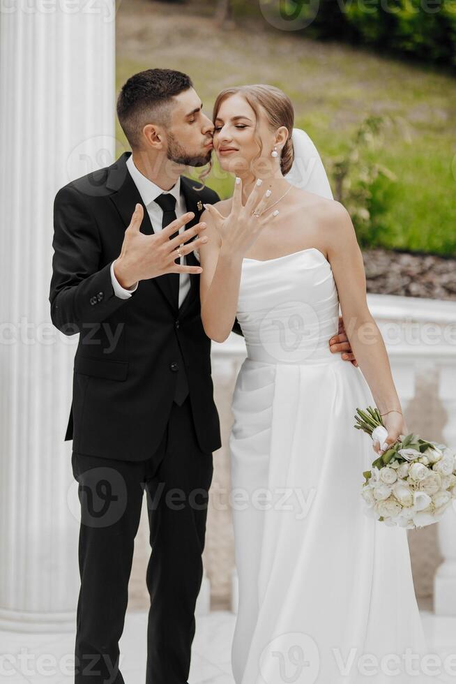 A bride and groom are kissing each other's ring fingers. The bride is wearing a white dress and the groom is wearing a black suit photo