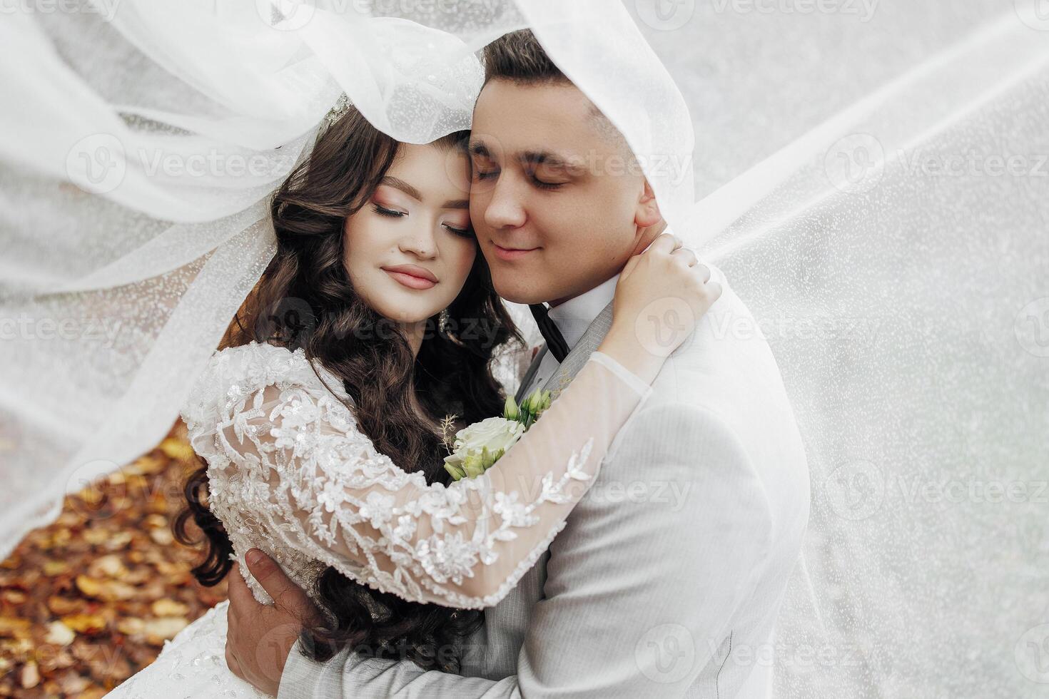 A bride and groom are hugging each other under a white veil. The bride is wearing a long dress and the groom is wearing a suit. Scene is romantic and happy photo