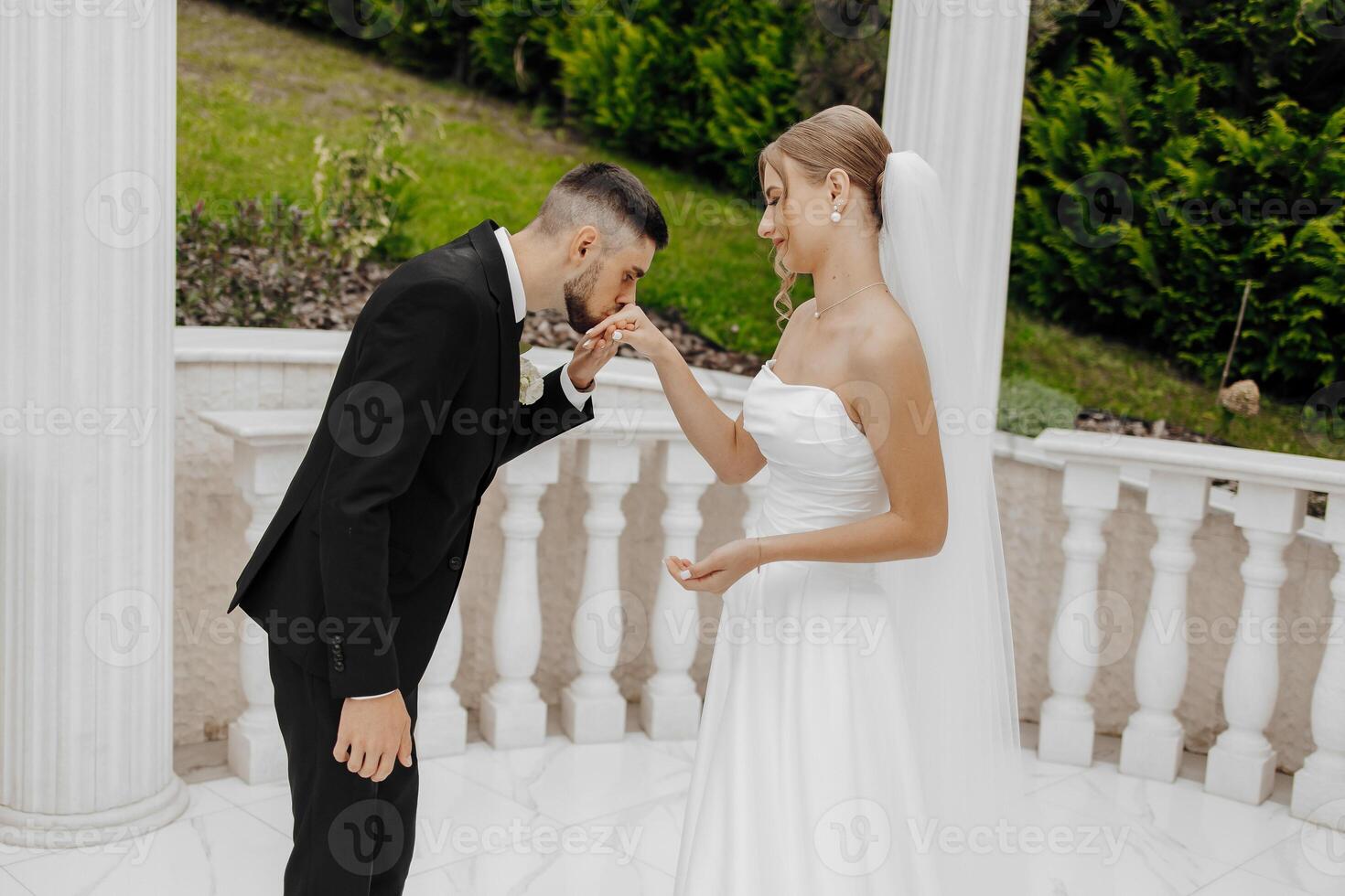A bride and groom are standing on a white marble balcony, the bride is wearing a white dress and the groom is wearing a black suit photo
