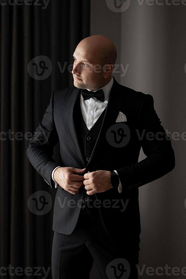 A man in a black suit and white shirt is getting ready to go out. He is adjusting his tie and looking at the camera photo