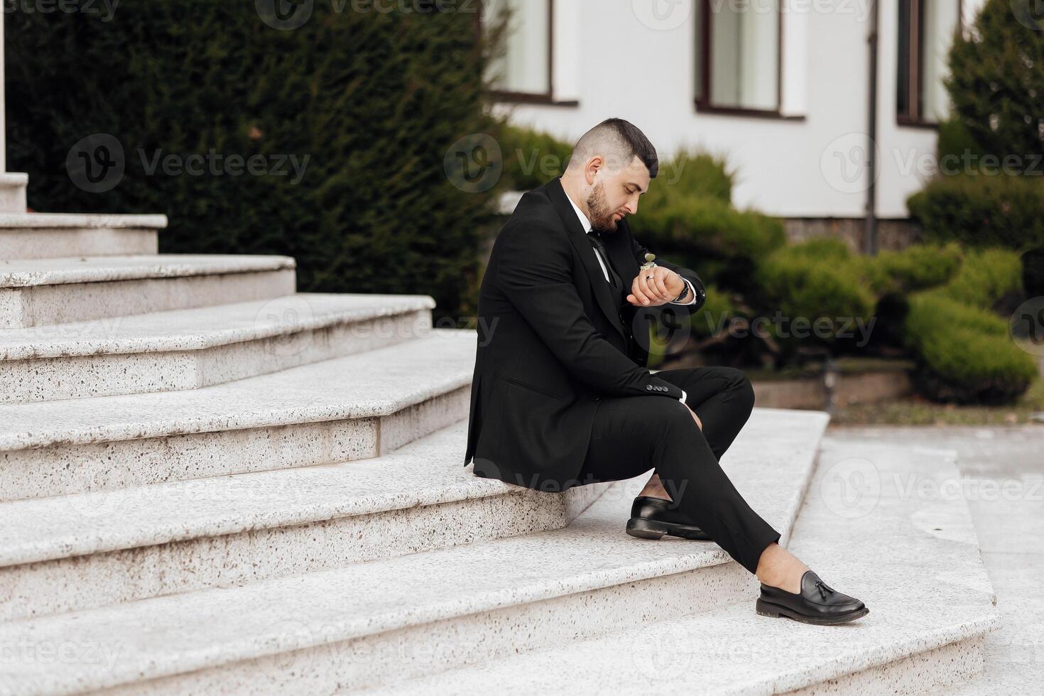 A man in a suit is sitting on a set of stairs. He is wearing a watch and he is checking the time. Concept of formality and professionalism, as the man is dressed in a suit and tie photo