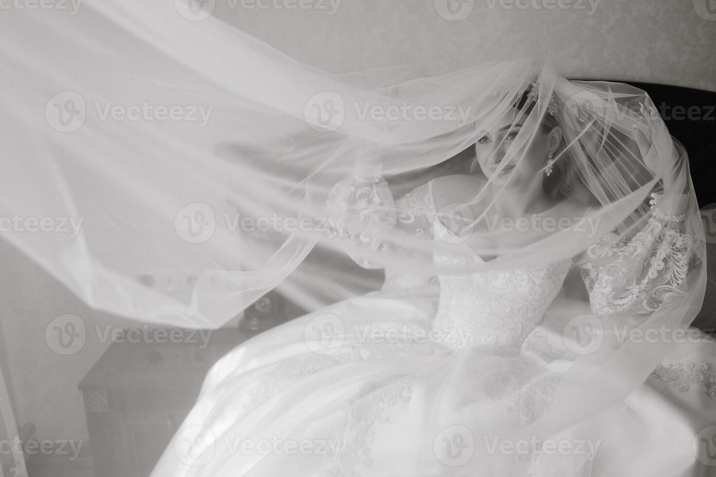 A bride is getting ready for her wedding and is wearing a white dress. The dress is flowing and has a veil on it. The bride is standing in front of a bed, and the bed is covered with a white sheet photo