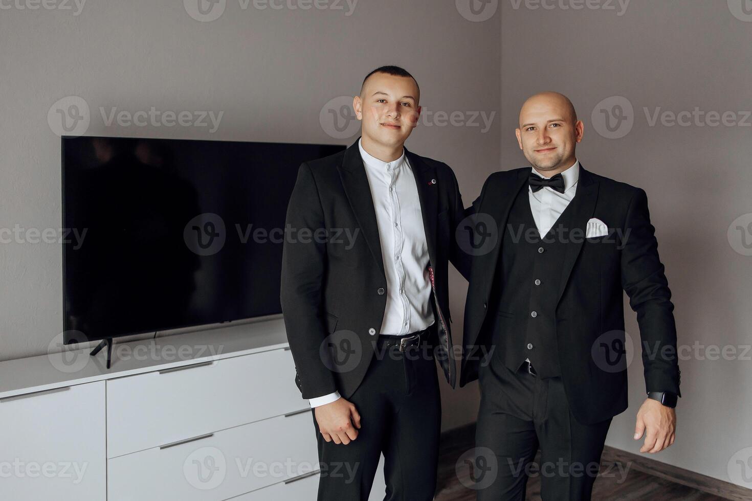 Two men in suits are standing next to each other in a room. One of them is wearing a tie photo