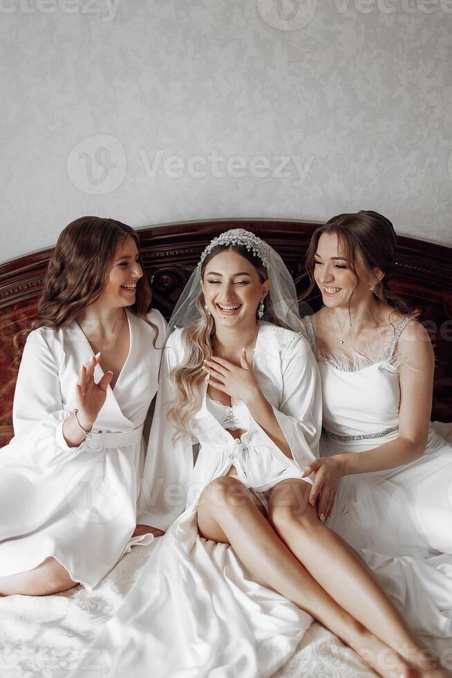 Three women are sitting on a bed, one of them is wearing a wedding dress. They are smiling and seem to be enjoying each other's company. Scene is happy and friendly photo