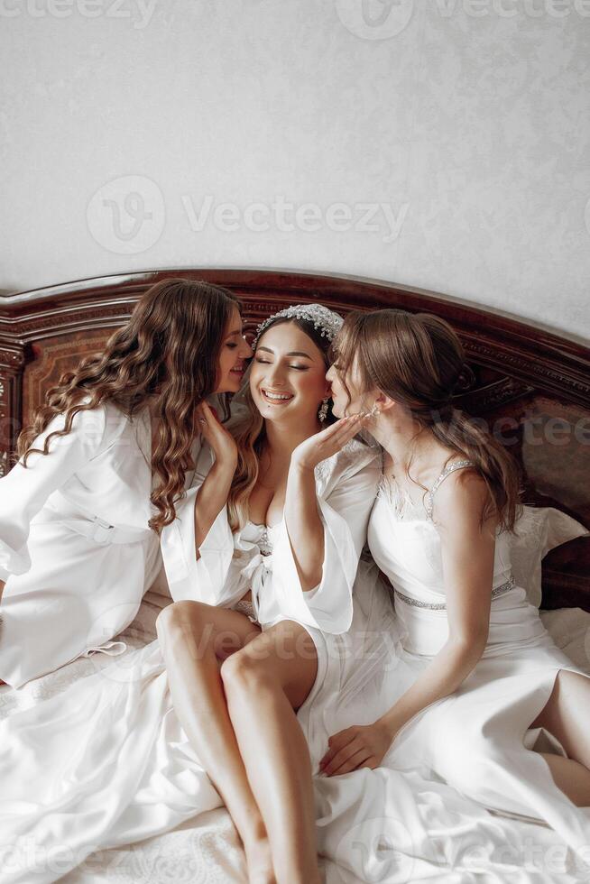 Three women are sitting on a bed, one of them is wearing a wedding dress. They are hugging and kissing each other, which suggests a close relationship between them. Scene is warm and affectionate photo