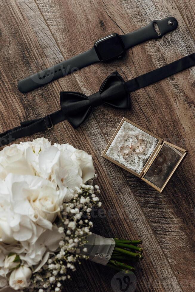 A bouquet of white flowers sits on a wooden table next to a watch and a tie. The flowers are arranged in a vase and the tie is folded photo