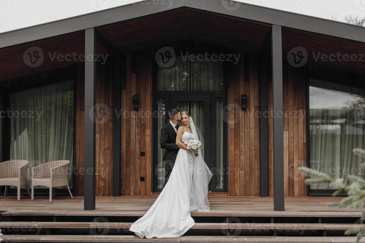 A bride and groom stand on a wooden deck, holding hands and smiling. The bride is wearing a white dress and the groom is wearing a suit. They are surrounded by chairs and a potted plant photo