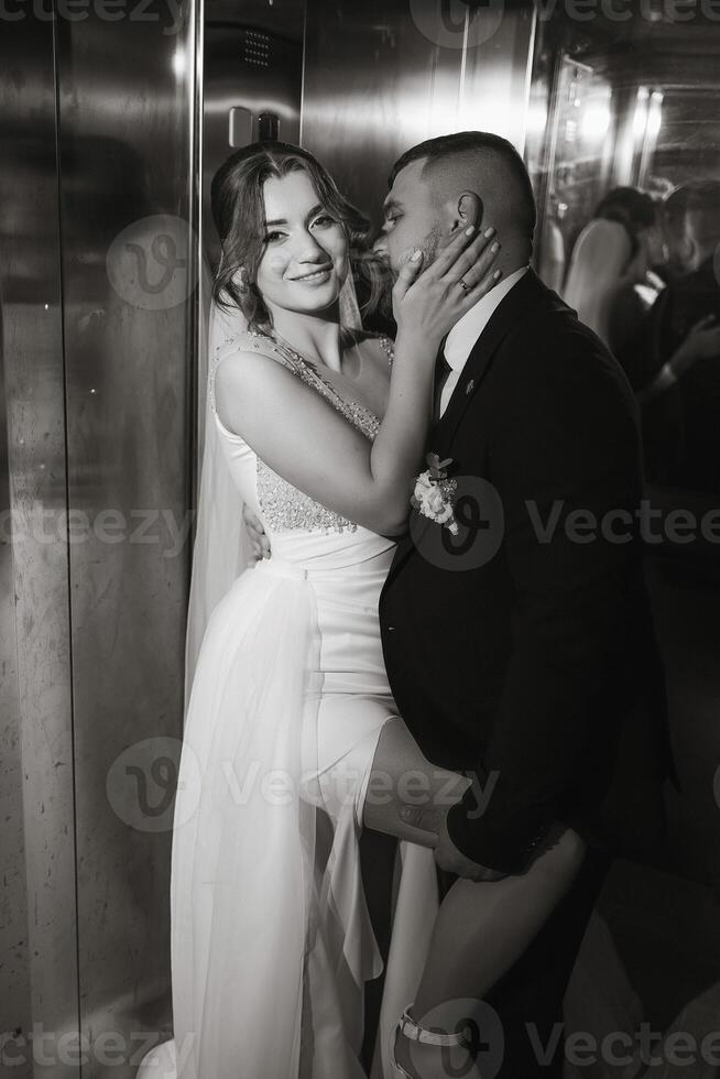 A bride and groom are kissing in an elevator. The bride is wearing a white dress and the groom is wearing a suit photo
