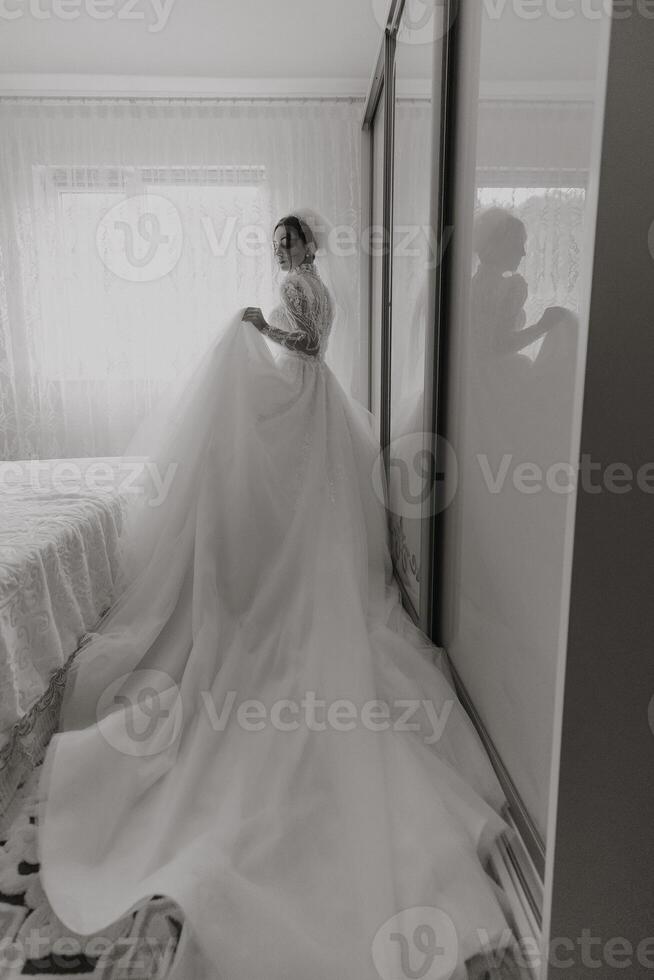 A woman in a wedding dress is standing in front of a mirror. The dress is long and flowing, and the woman is holding a veil. The image has a romantic and elegant mood photo