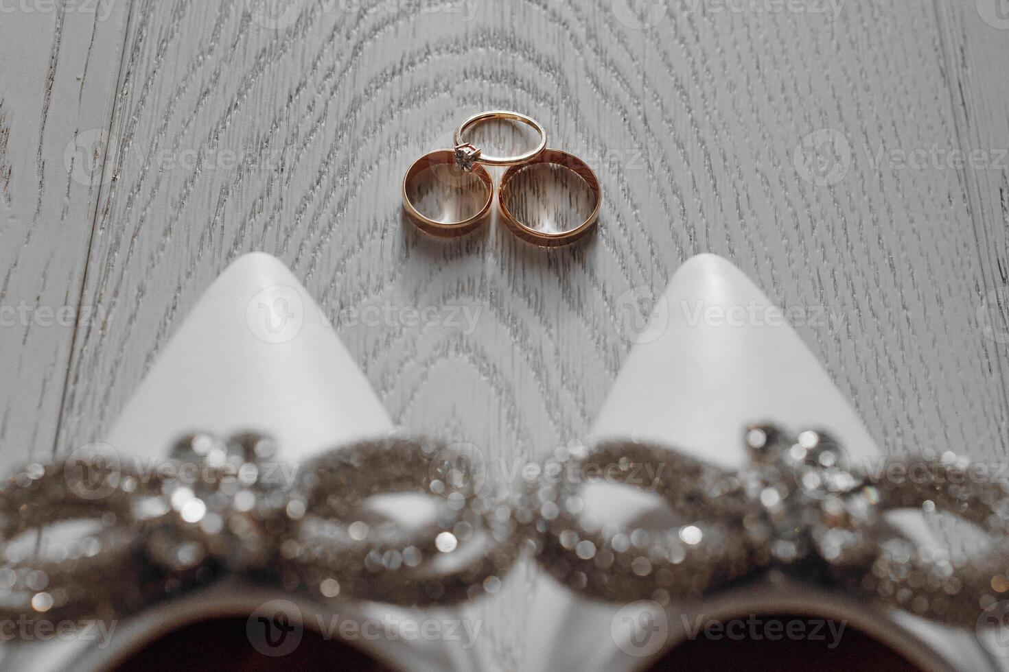 A pair of white shoes with a bow on the front and a ring on the toe. The shoes are next to two gold wedding rings photo