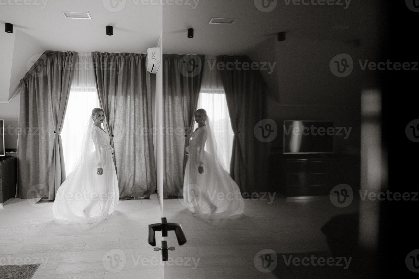 A woman in a wedding dress is standing in front of a mirror. The room is dimly lit, and the curtains are drawn, creating a sense of intimacy and privacy. The woman is posing for a picture photo