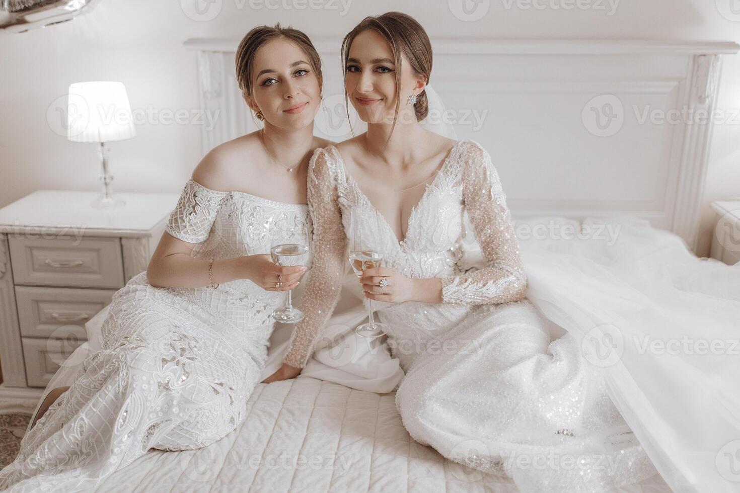 Two women in wedding dresses are sitting on a bed, holding glasses of wine. Scene is lighthearted and celebratory, as the women are enjoying each other's company and the special occasion photo
