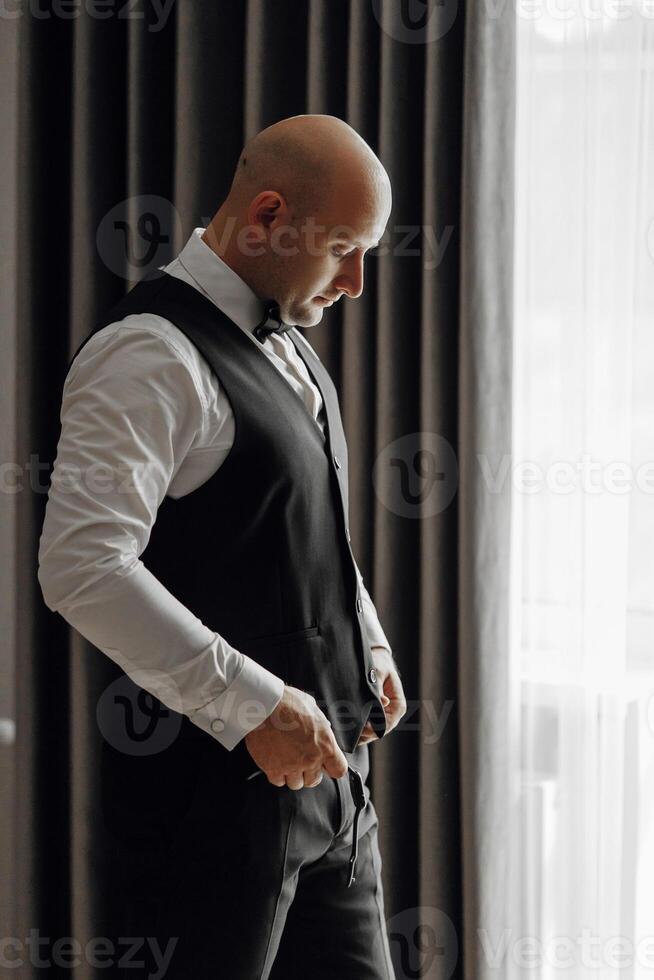 A man in a black vest and white shirt is standing in front of a window. He is adjusting his pants and looking out the window. Concept of formality and attention to detail photo