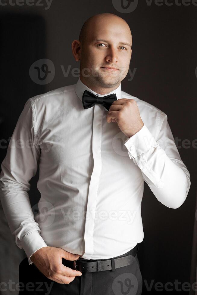 A man in a white shirt and black bow tie is standing in front of a wall. He is adjusting his tie and looking at the camera photo